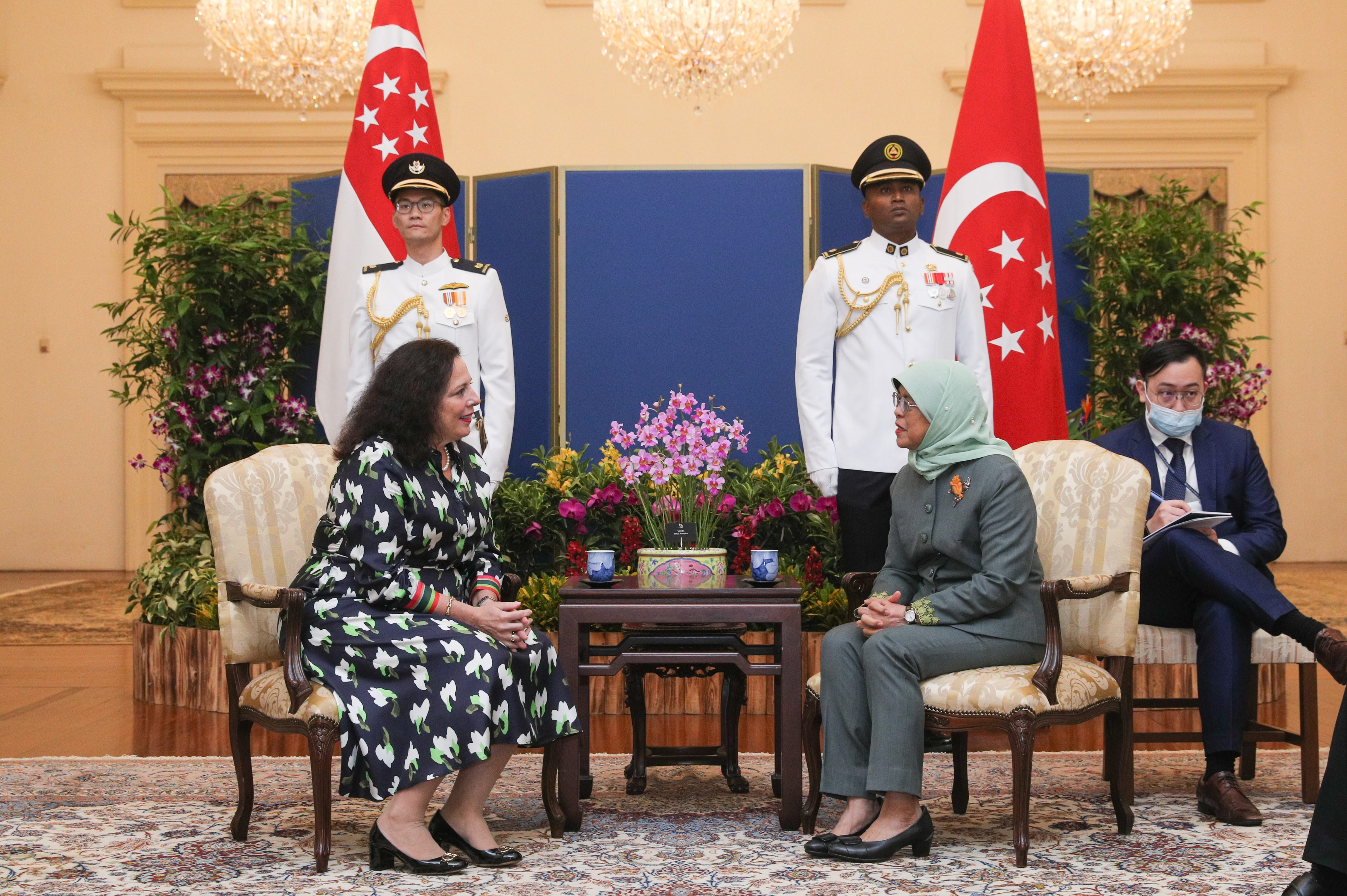 Two women sit in chairs flanked by Singapore flags and uniformed guards in an ornate room.